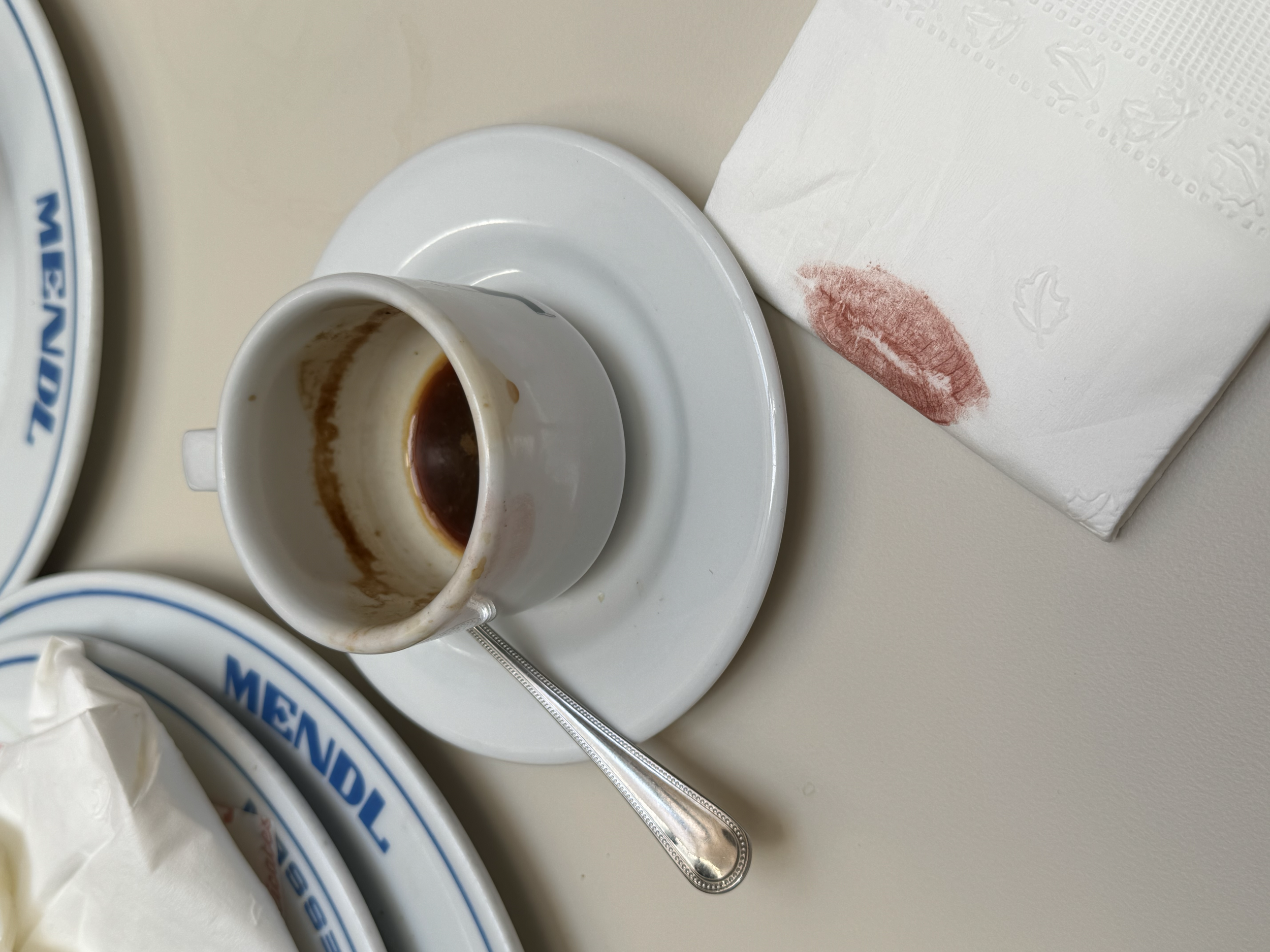 A white coffee cup with used coffee inside, on a white saucer, with a teaspoon, porcupine kiss mark on a white napkin, and part of a blue and white plate that says 'MENDL' in the background.