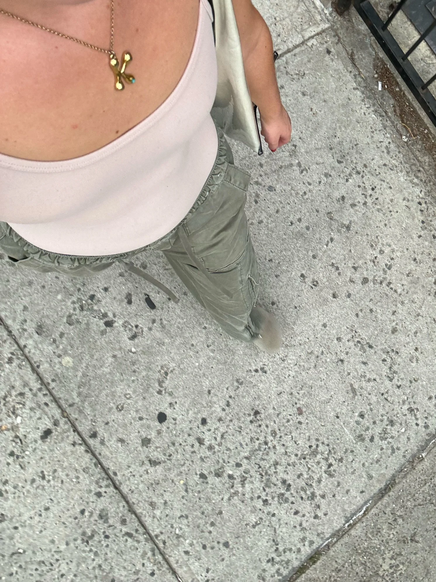 A top-down view of a person standing on a concrete sidewalk, wearing a light-colored tank top, cargo pants, and a necklace with a butterfly pendant. The person's right hand holds a white bag, and part of their arm and hand are visible.