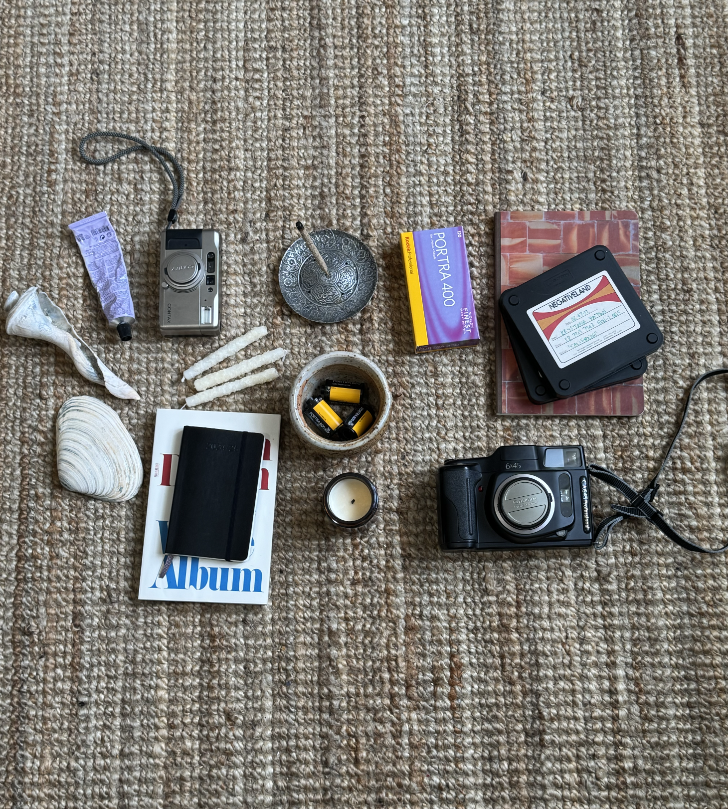 A collection of personal items on a beige carpet, including a vintage camera, a compact digital camera, a pack of Kodak Portra 400 film, a notebook, batteries, seashells, a small candle, and various decorative objects.