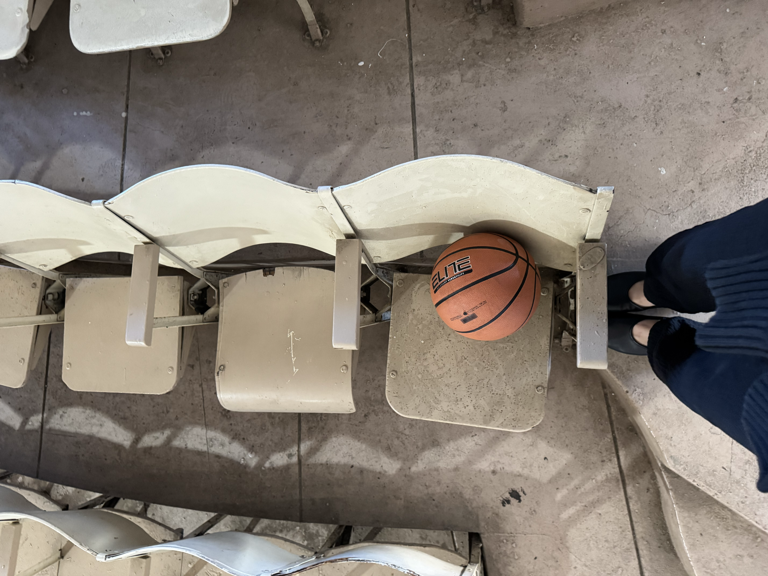 A row of empty beige stadium seats with a basketball on one of the seats, and a pair of people's legs in black shoes and dark pants standing nearby.