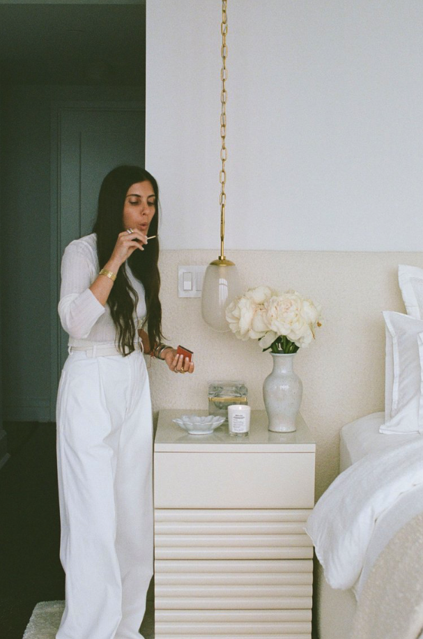 A woman with long dark hair wearing white high-waisted pants and a white top, standing next to a bedside table with a vase of white flowers, a candle, and a small dish, in a bedroom with a white headboard, white bedding, and a wall-mounted lamp.