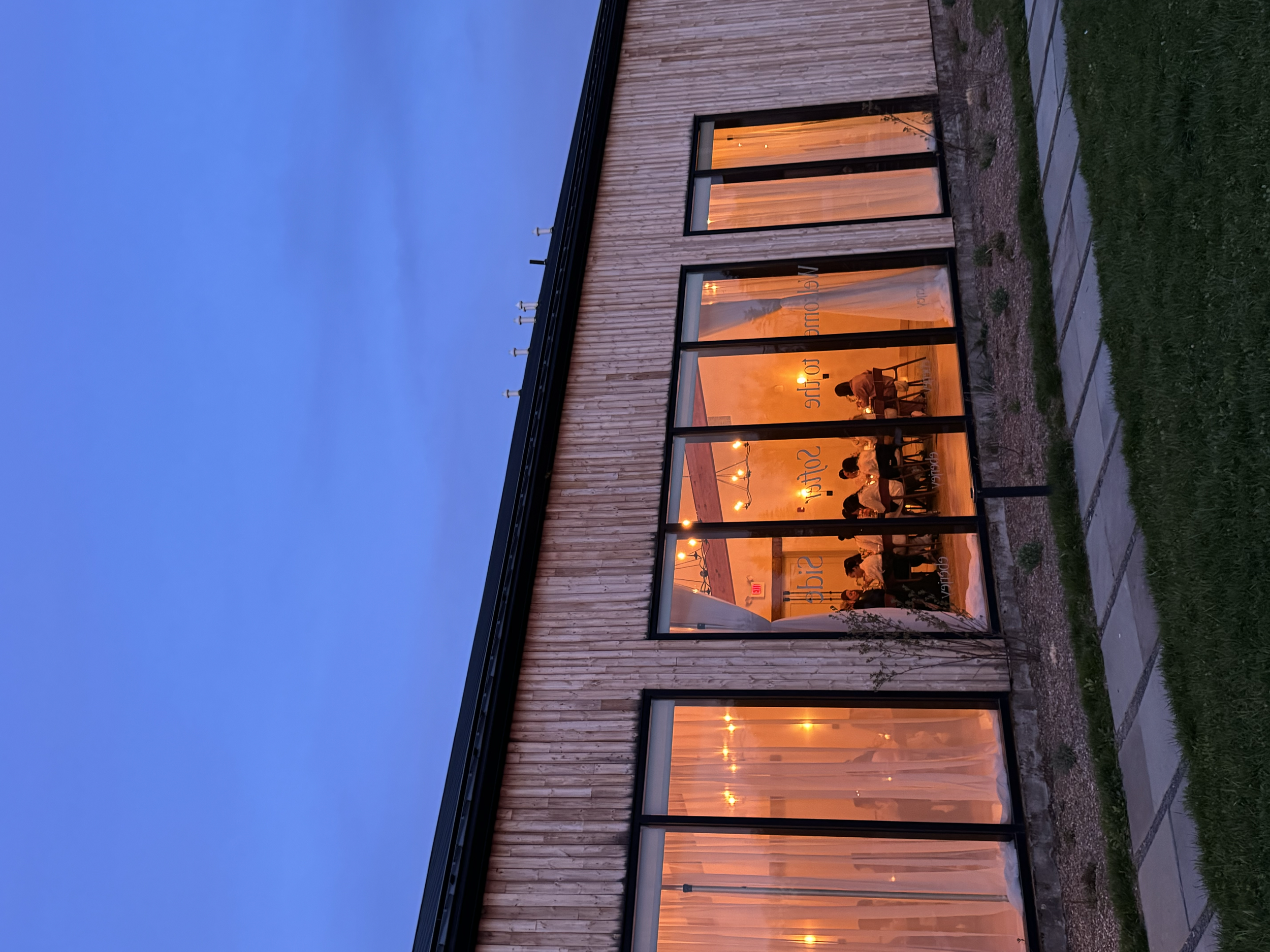 The interior of a cozy restaurant seen through large glass windows, with warm lighting, seating areas, and a few patrons dining inside during dusk.