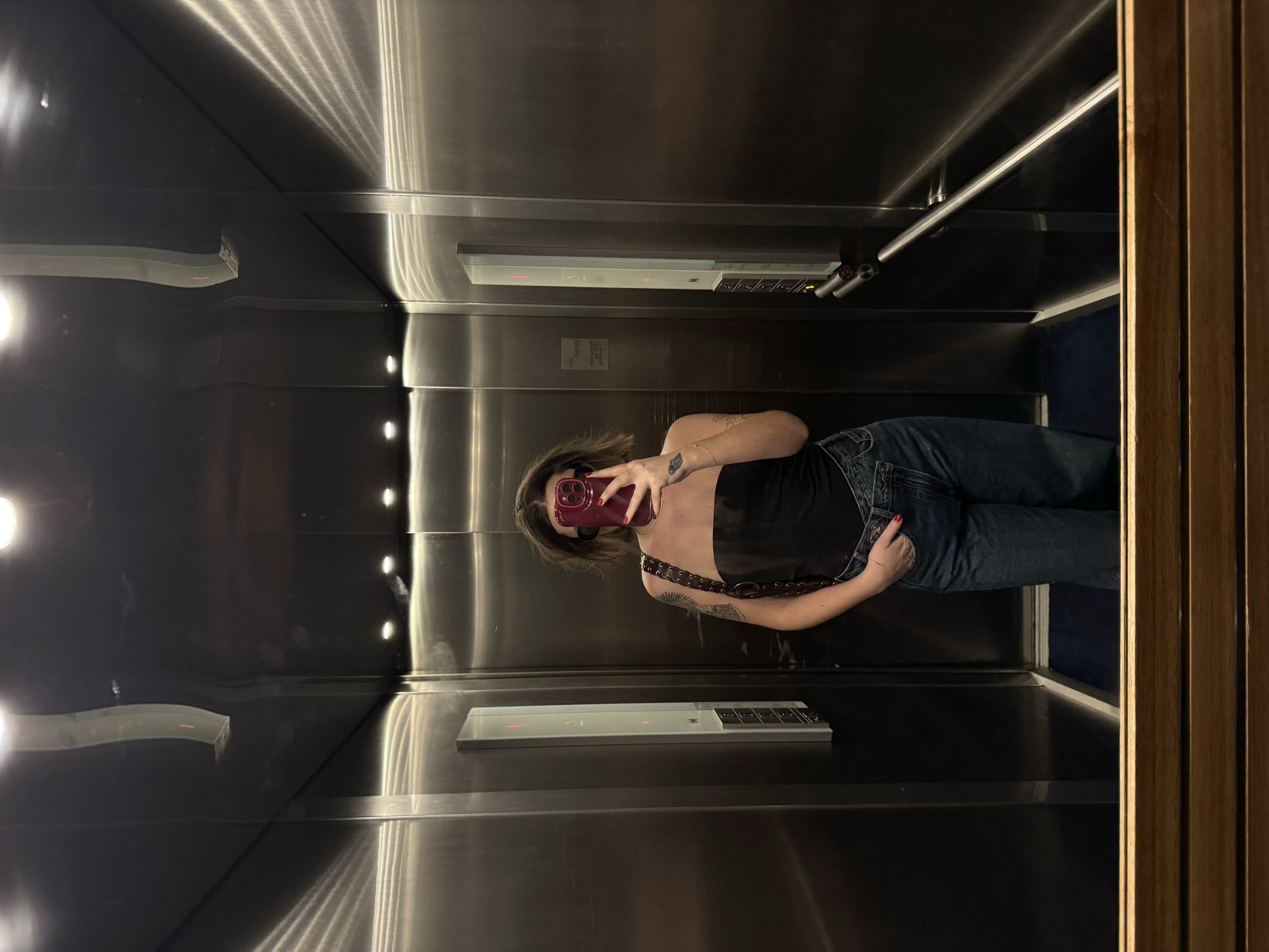 A person taking a mirror selfie inside an elevator with stainless steel walls, wearing a black strapless top and jeans, holding a pink phone covering their face.
