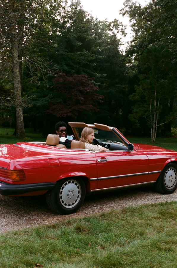Two women riding in a red convertible car parked on grass with trees in the background