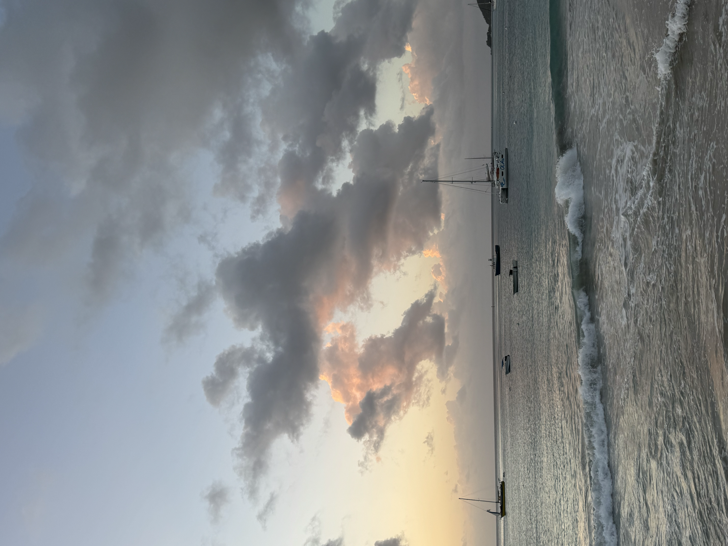 Sunset over a calm ocean with clouds in the sky, sailboats floating on the water, waves gently hitting the shore.