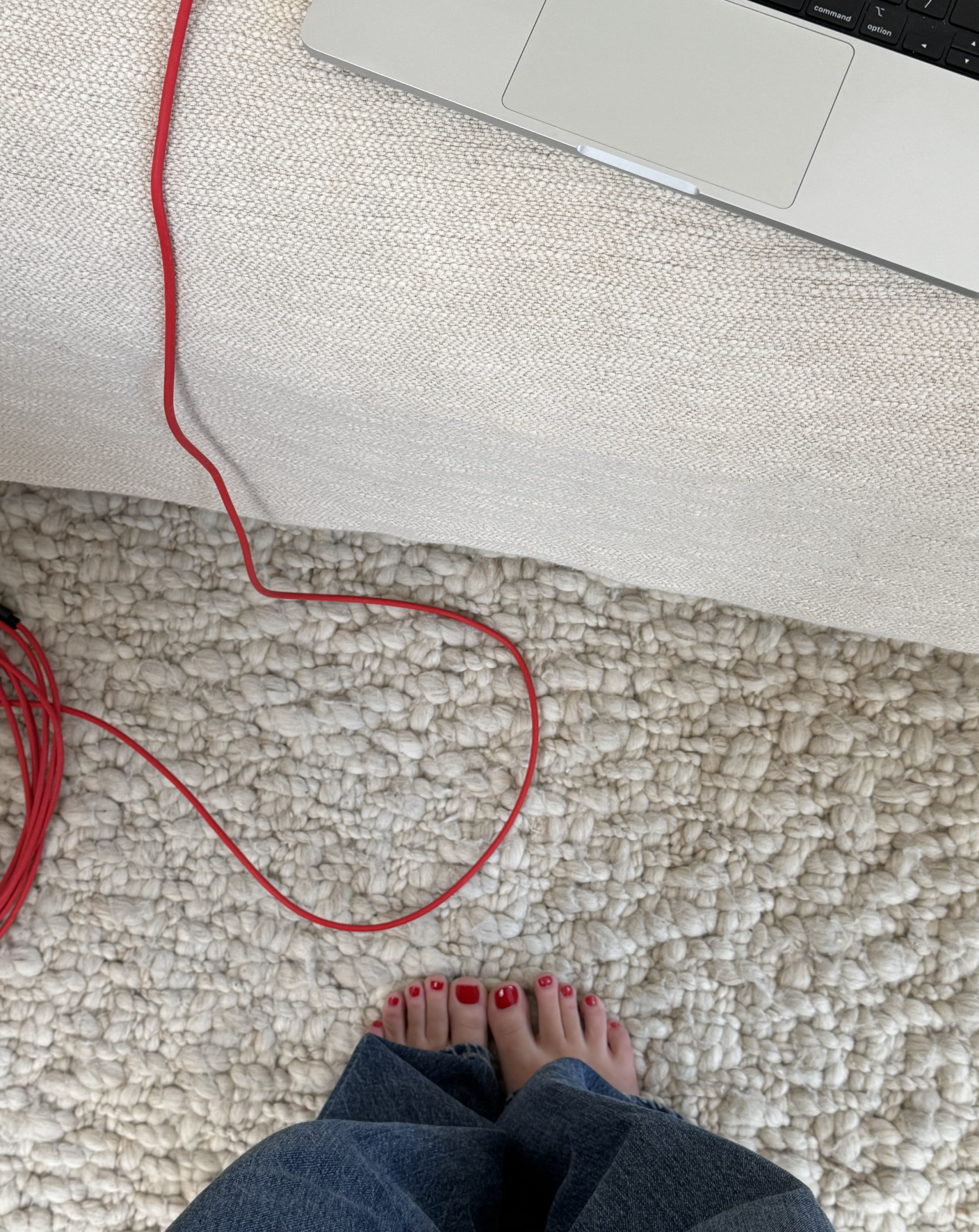 Person standing on a plush carpet with red painted toenails, near a beige sofa and a silver laptop with red charging cable.