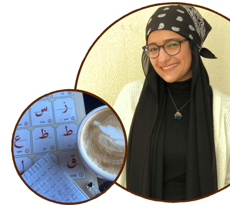 A woman smiling, wearing glasses, a bandana, and a black scarf, with a coffee and Arabic alphabet learning materials.