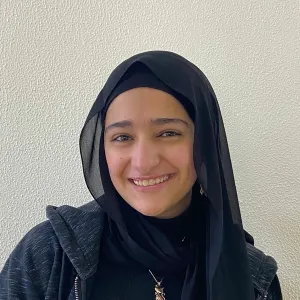 A young woman wearing a black headscarf and a dark jacket, smiling at the camera, against a plain light-colored wall.