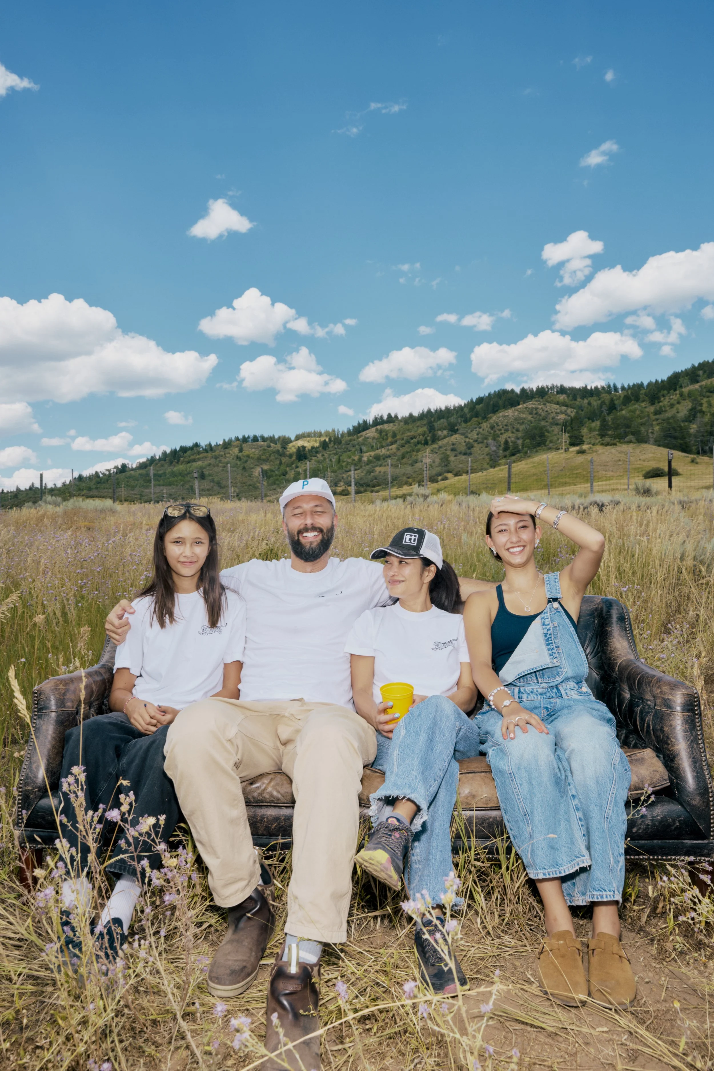 A group of four people sitting on a vintage couch outdoors in a grassy field with mountains and a blue sky with clouds in the background.