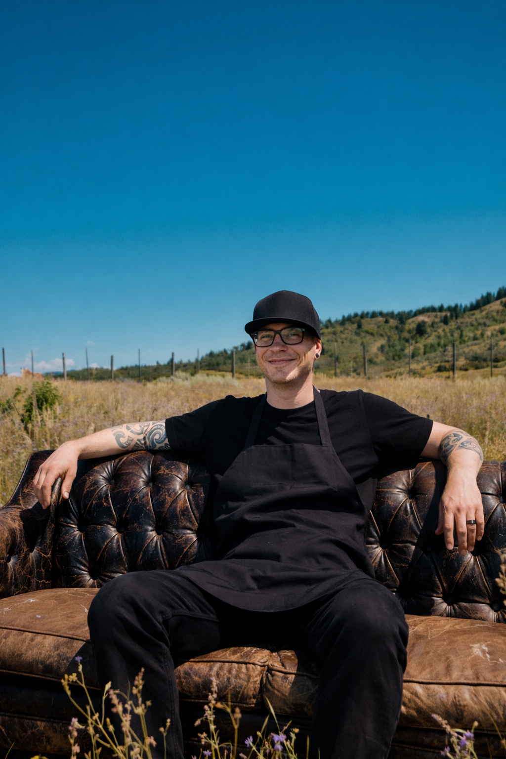 A young man with tattoos, wearing glasses, a black hat, a black t-shirt, and a black apron, sitting on a vintage brown leather couch outdoors on a sunny day with a rural landscape and clear blue sky in the background.