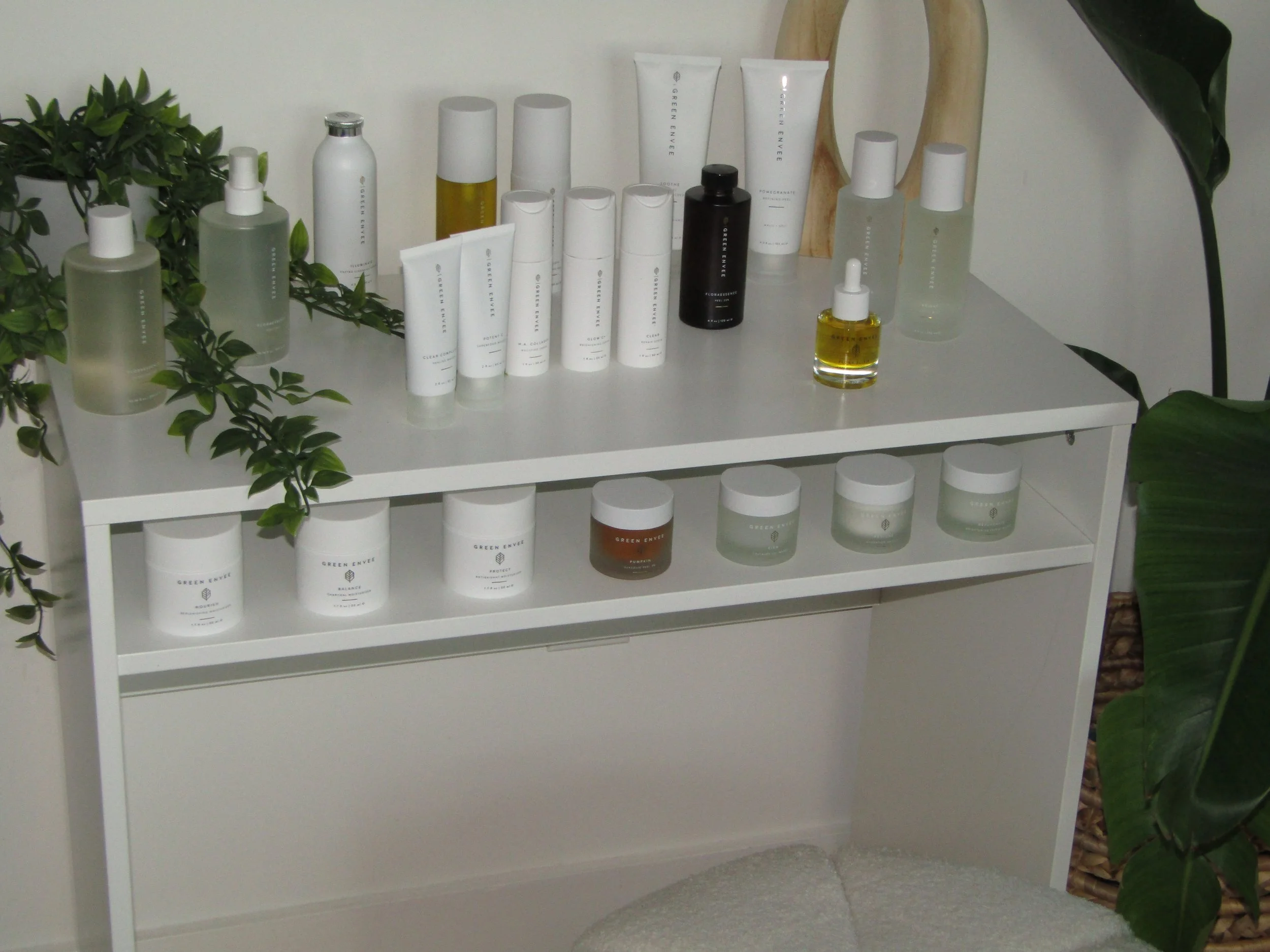 A white shelf displaying various skincare products including creams, serums, and oils, surrounded by green plants.