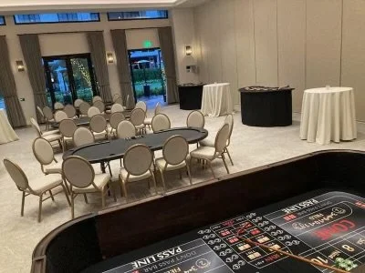 Casino-themed event room with roulette and card tables, round banquet tables with chairs, and white tablecloths, set in a spacious area with large windows and neutral decor.