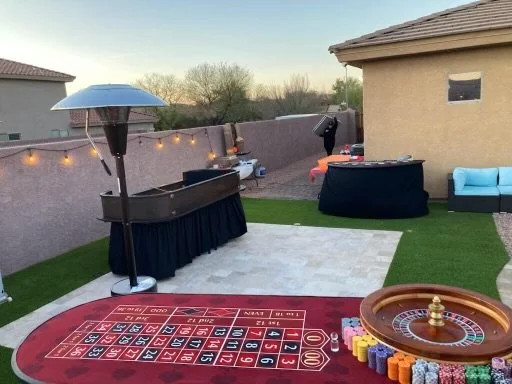Outdoor backyard setup with casino theme, featuring a red roulette table, a craps table, patio heater, and outdoor string lights on a patio with grass and stone flooring.