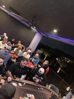 A group of people playing poker at a card table outdoors at night, with palm trees visible in the background and dim overhead lighting.