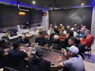 A room filled with people playing poker around two tables, under neon lighting. Players are engaged in gameplay, chips and cards are visible. Casual attire, including baseball caps and sports jerseys, is worn by participants.