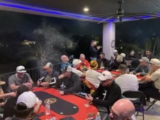 Group of people playing poker on a covered patio at night with visible smoke and neon lighting.