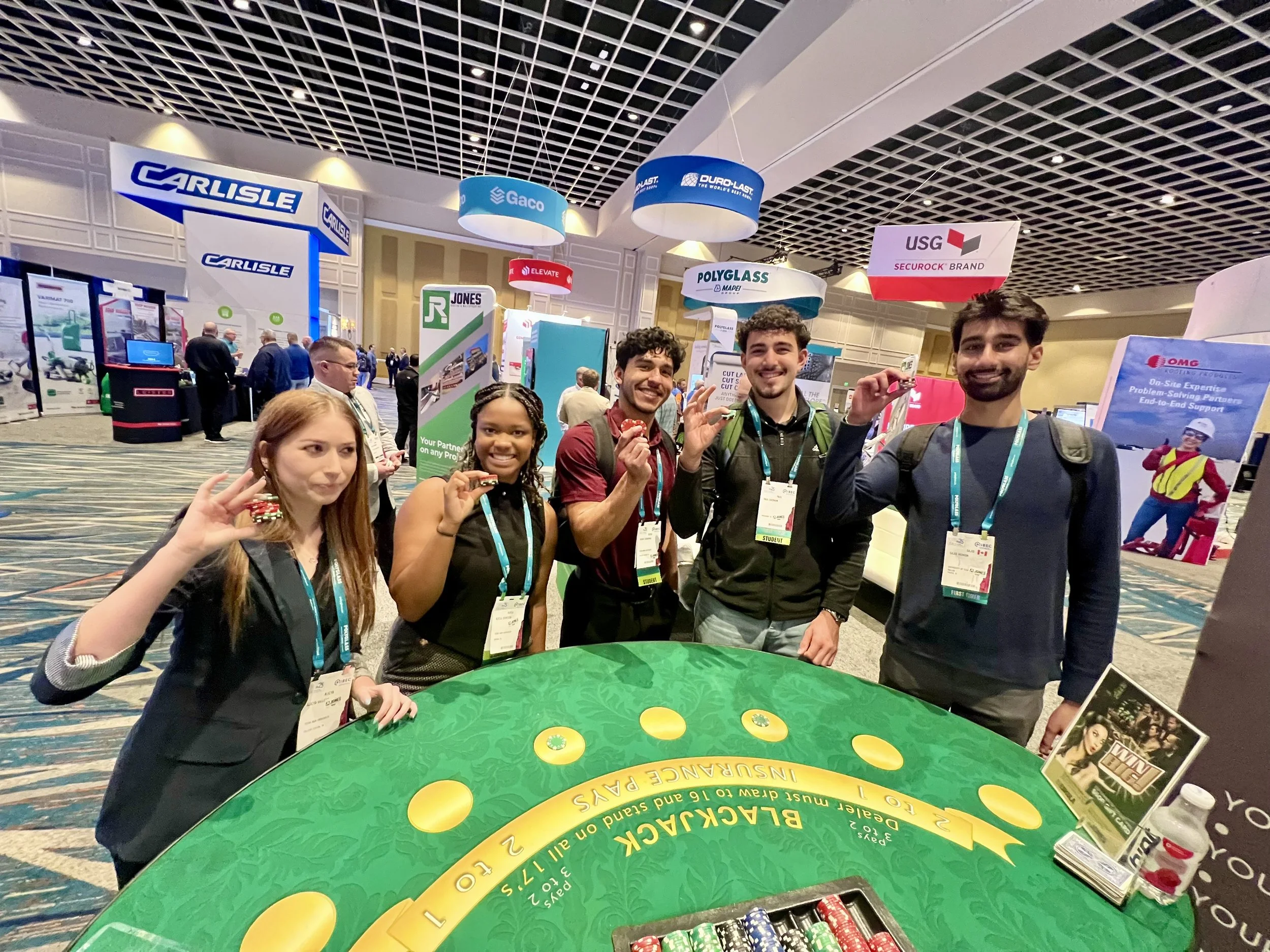 Group of five people standing around a blackjack table at a trade show, holding poker chips. They are smiling and wearing event badges. Various company banners and booths are visible in the background.