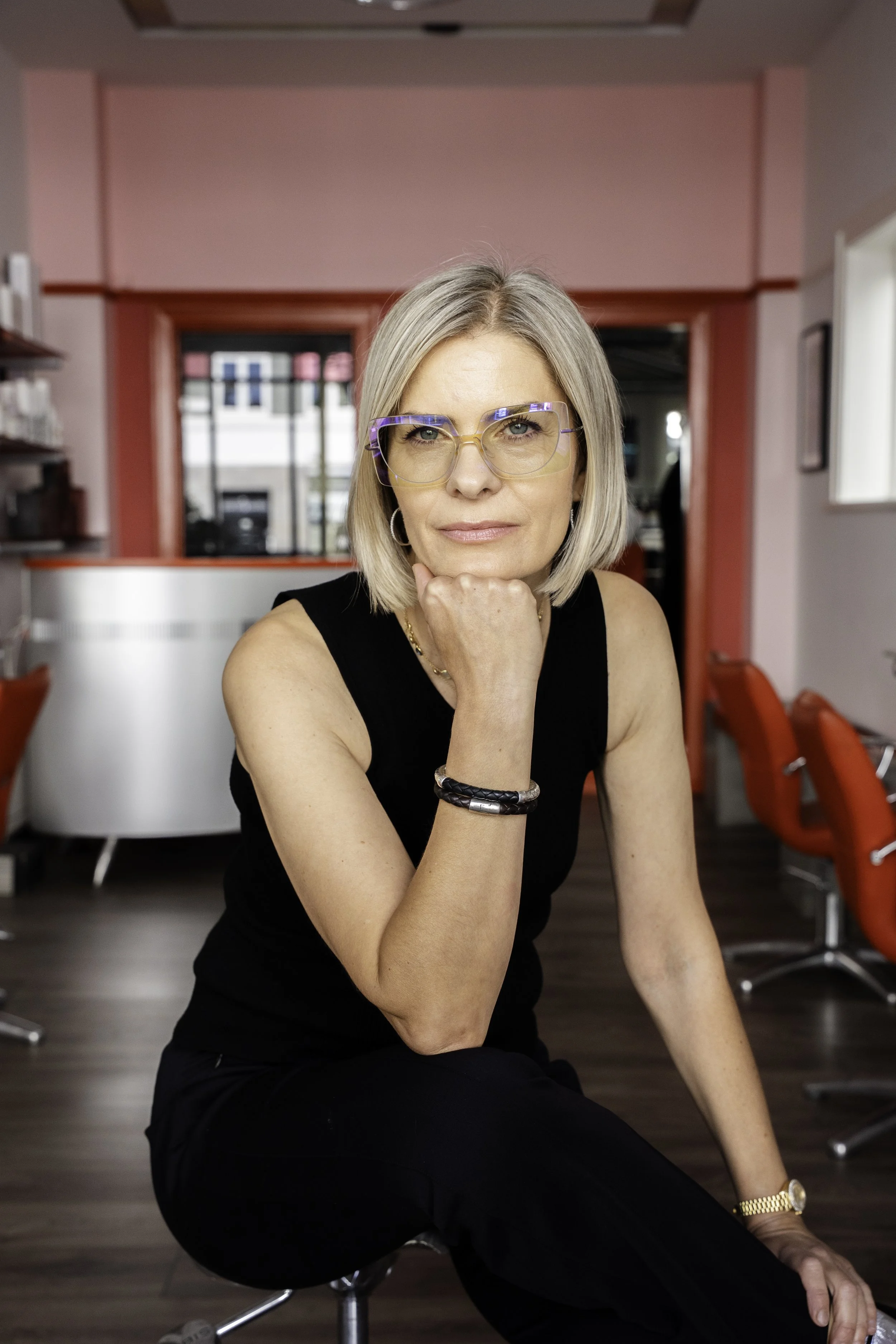 En kvinde med blondt hår, iført briller, en sort ærmeløs top og sorte bukser, siddende i en salon med røde stole og lyserøde vægge.