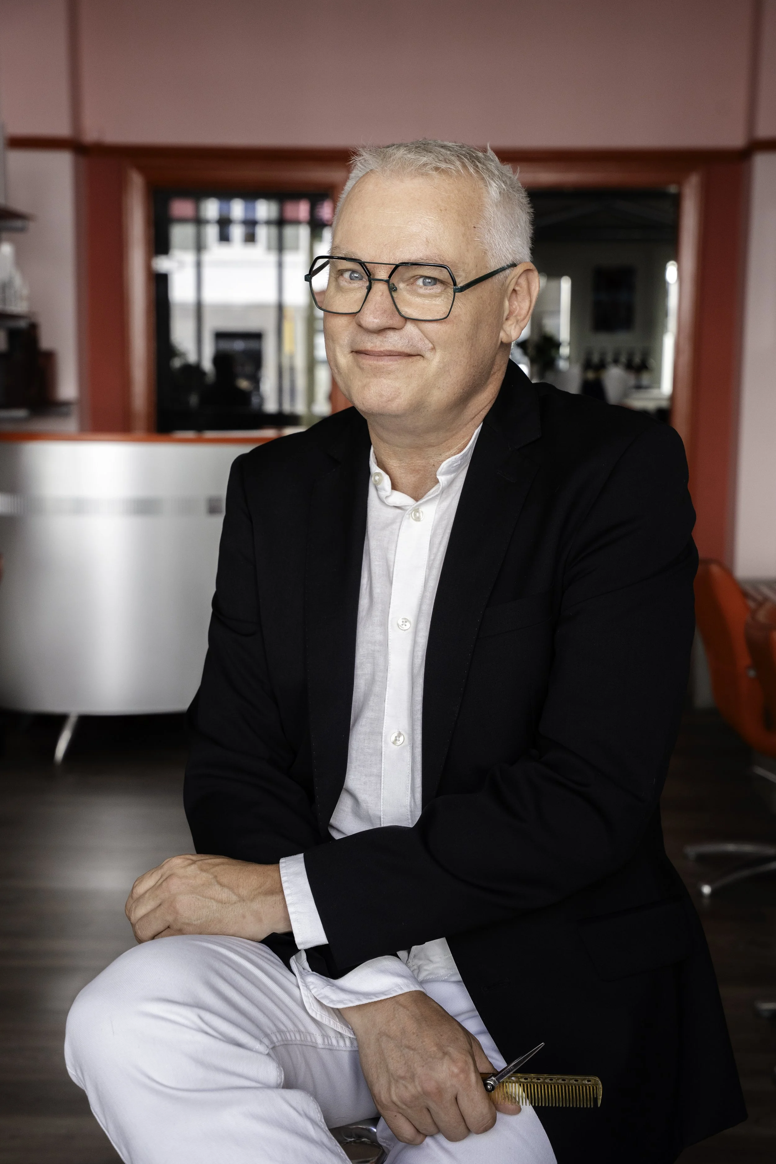 En midaldrende mand med lyst gråt hår og briller, iført en hvid skjorte og sort blazer, sidder indendørs med et smil og holder en kam i højre hånd i et retroinspireret rum med pink og røde detaljer.