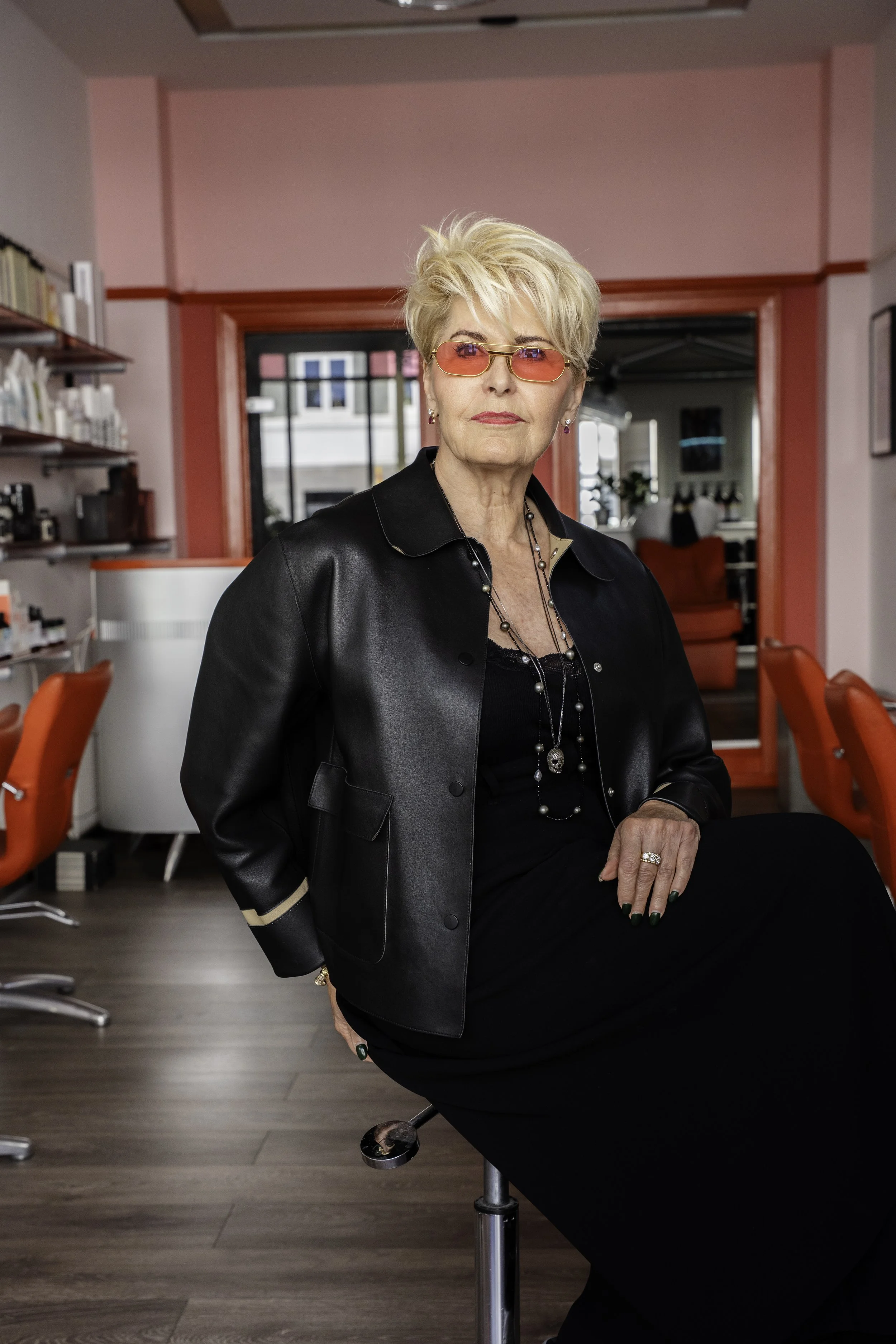 En ældre kvinde med kort blondt hår, iført lyserøde, tonede briller, en sort læderjakke og en sort kjole, siddende på en stol i et farverigt rum med røde stole i frisørsalon og udstyr samt lyserøde vægge.