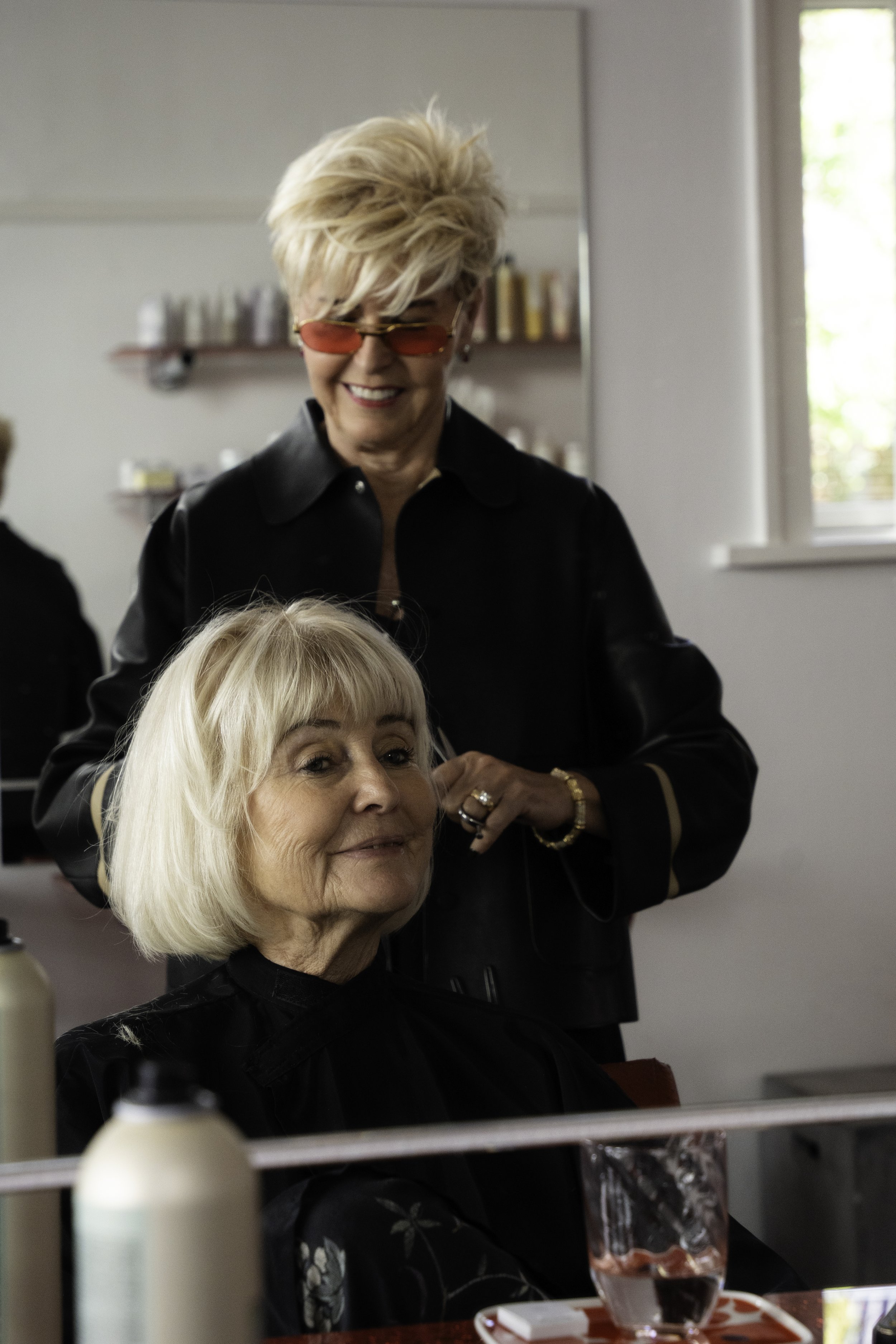 En frisør iført sort læderjakke og orange solbriller smiler, mens han arbejder på en ældre kvindes hår i en salon. Den ældre kvinde med blondt hår og en blomstret bluse sidder smilende, med et glas vand og nogle hårstylingredskaber på bordet.