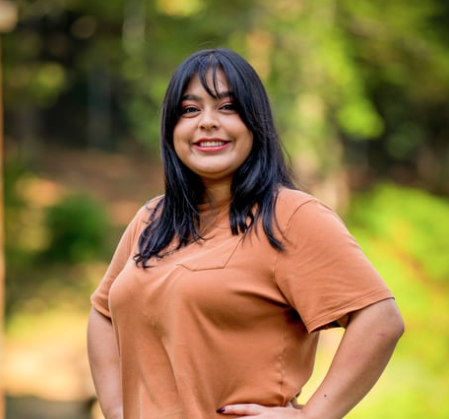 A woman smiling outdoors with green trees in the background.
