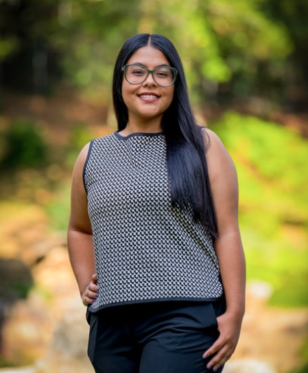 Young woman with long black hair and glasses standing outdoors in a green, blurred natural background.