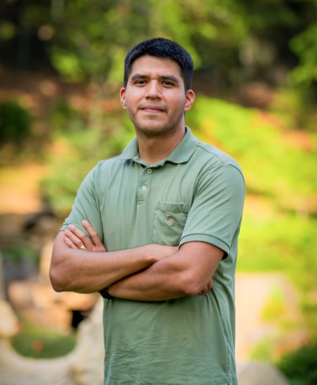Man with short black hair, wearing a light green polo shirt, standing outdoors with arms crossed, in front of a blurred background of trees and greenery.