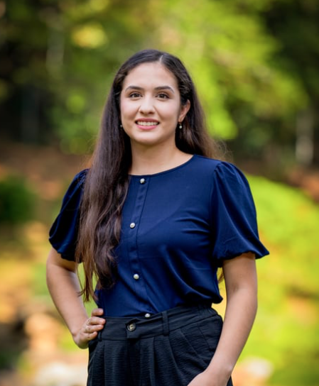 Young woman with long dark hair wearing a navy blouse and black pants standing outdoors with trees and greenery in the background.