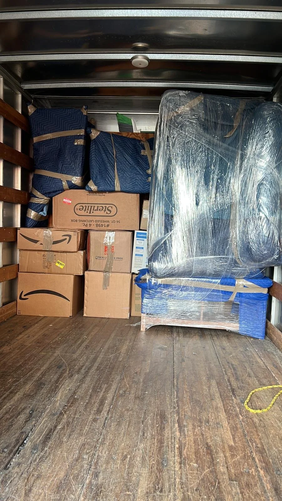 Modern house with wooden exterior in a field of tall dry grass, overlooking a lake with mountains in the background under a cloudy sky.