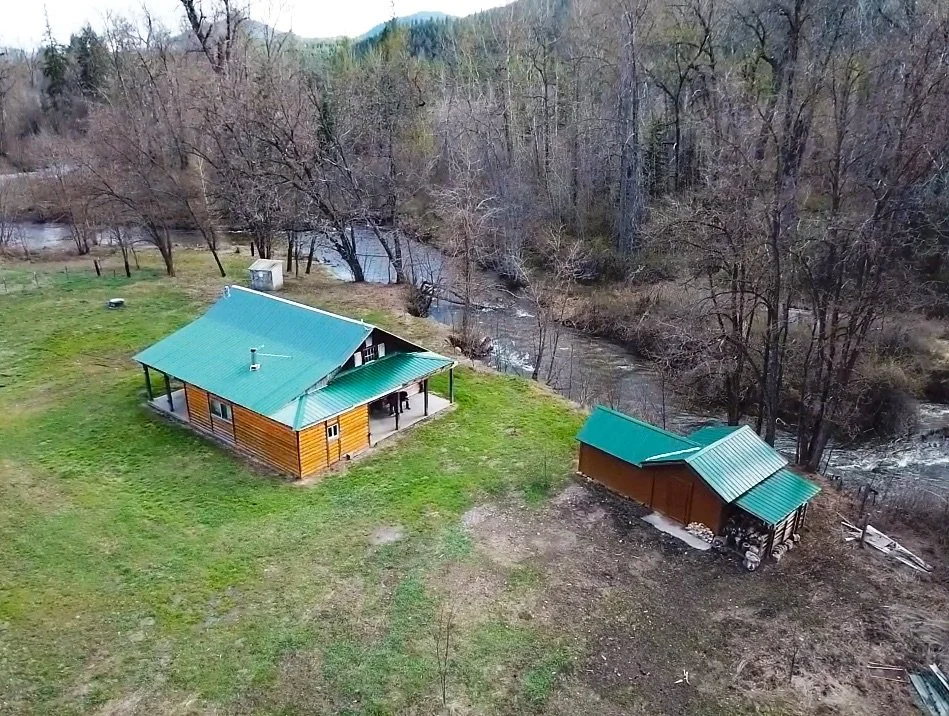 Aerial view of a rustic riverfront cabin with green metal roof, covered porch, and detached shed on open acreage, surrounded by trees and a flowing creek in a peaceful rural setting.
