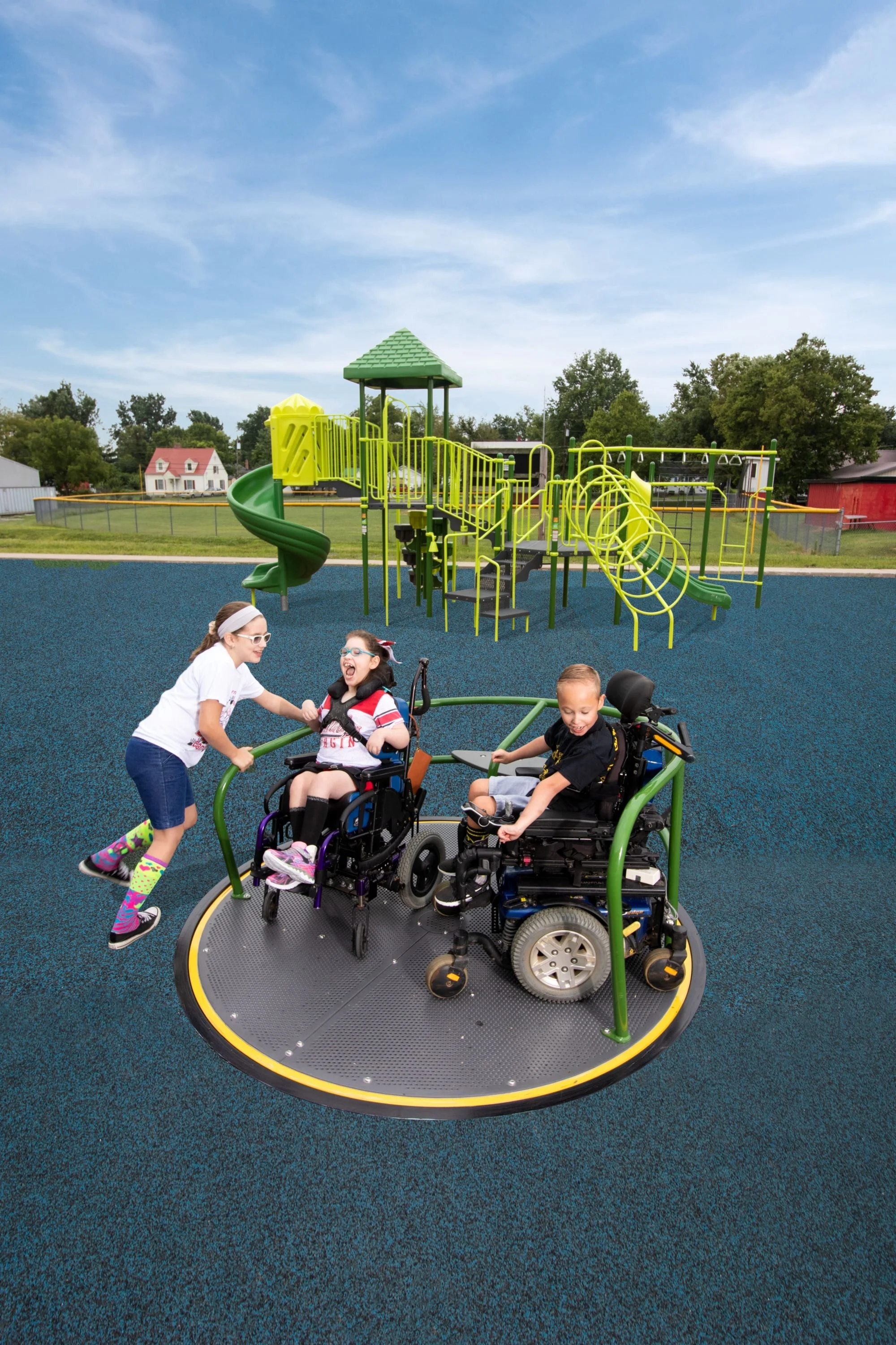 A playground with a blue rubber surface, a large climbing structure with tunnels and slides, and two children in wheelchairs being pushed on an in-ground merry-go-round, with a girl standing and pushing one of the children, under a partly cloudy sky.