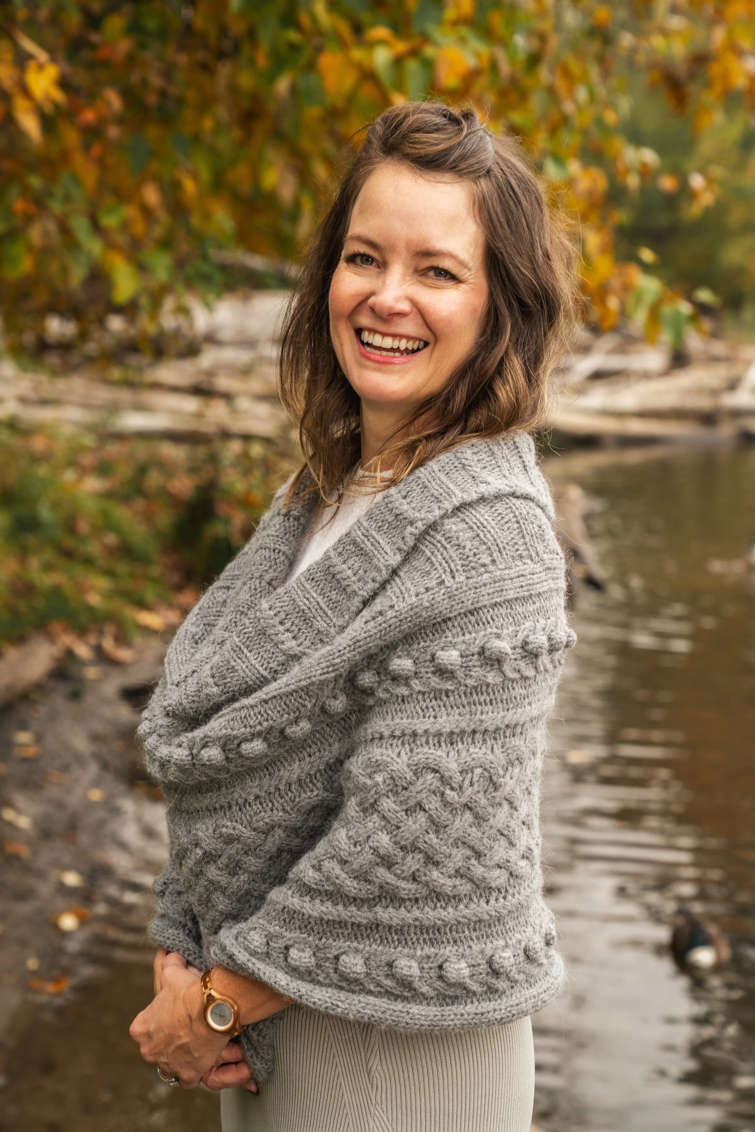 A woman with brown hair, smiling, wearing a gray knit sweater, standing outdoors near a creek with autumn leaves in the background.