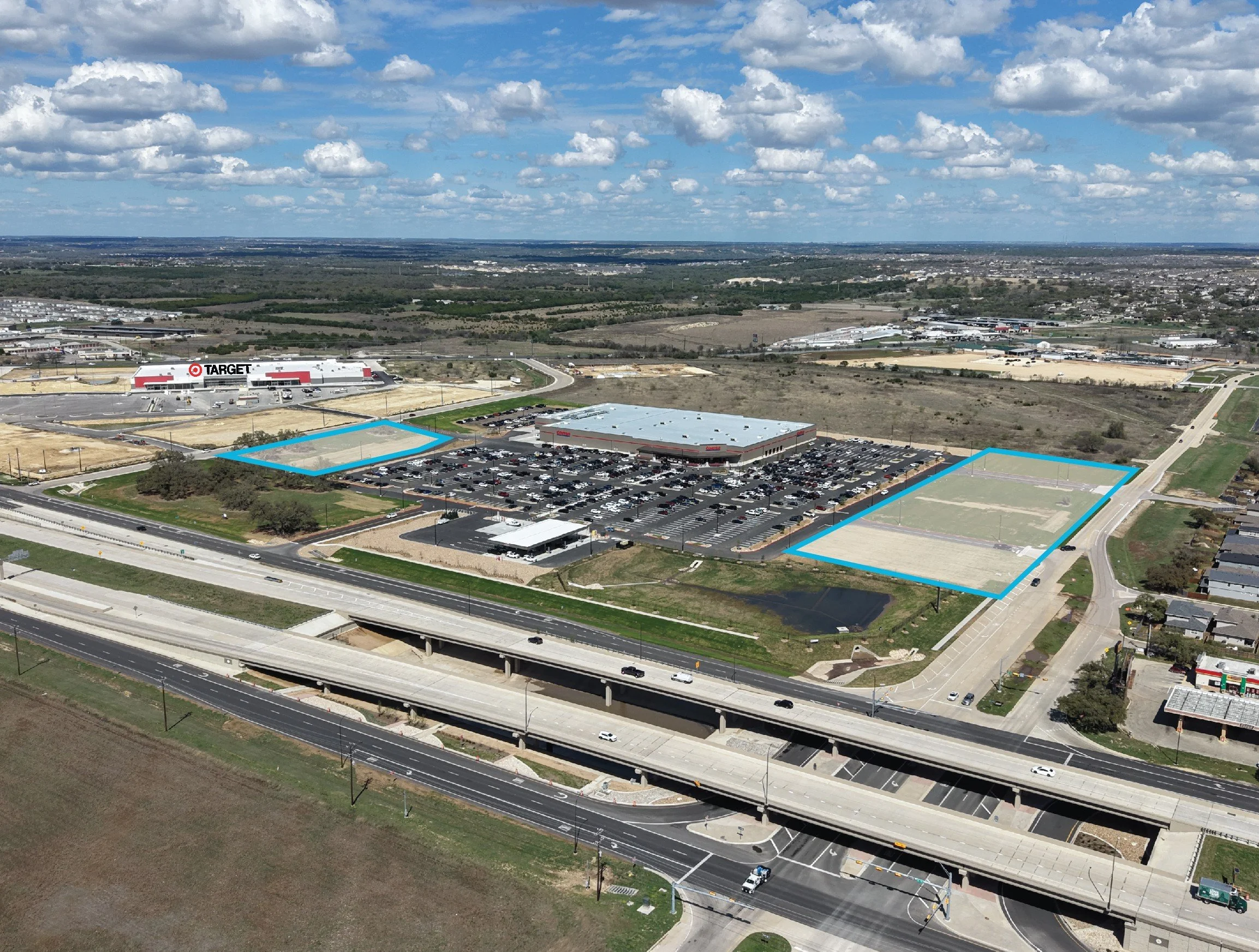 Aerial view of a shopping center parking lot with a Starbucks, Chipotle, and Chick-fil-A among trees and adjacent to a green area with tall office buildings in the background.