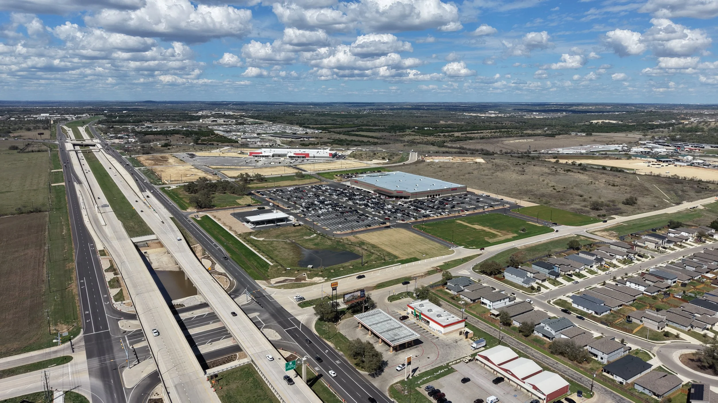 Aerial view of a shopping center parking lot with a Starbucks, Chipotle, and Chick-fil-A among trees and adjacent to a green area with tall office buildings in the background.
