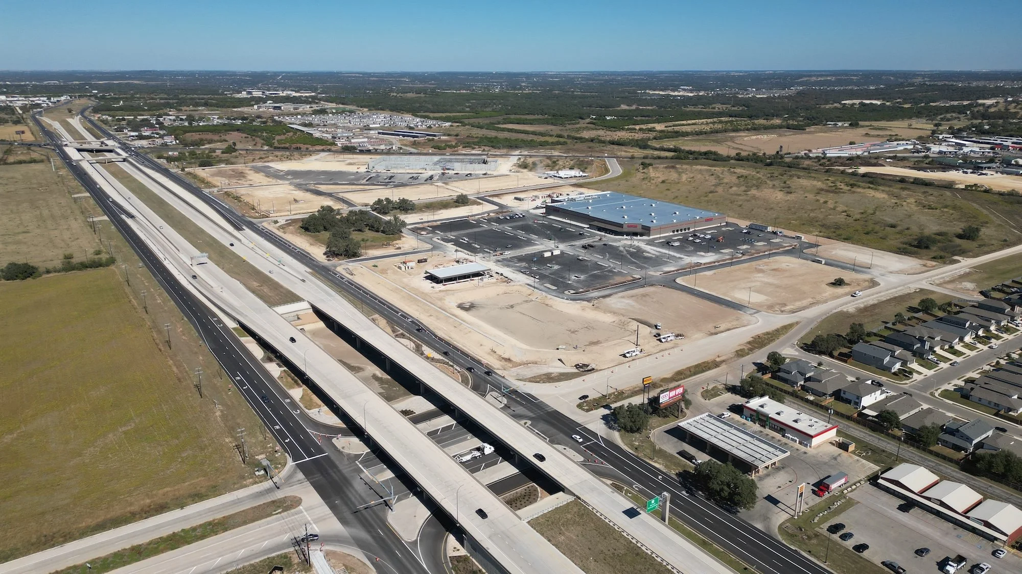 Aerial view of a shopping center parking lot with a Starbucks, Chipotle, and Chick-fil-A among trees and adjacent to a green area with tall office buildings in the background.