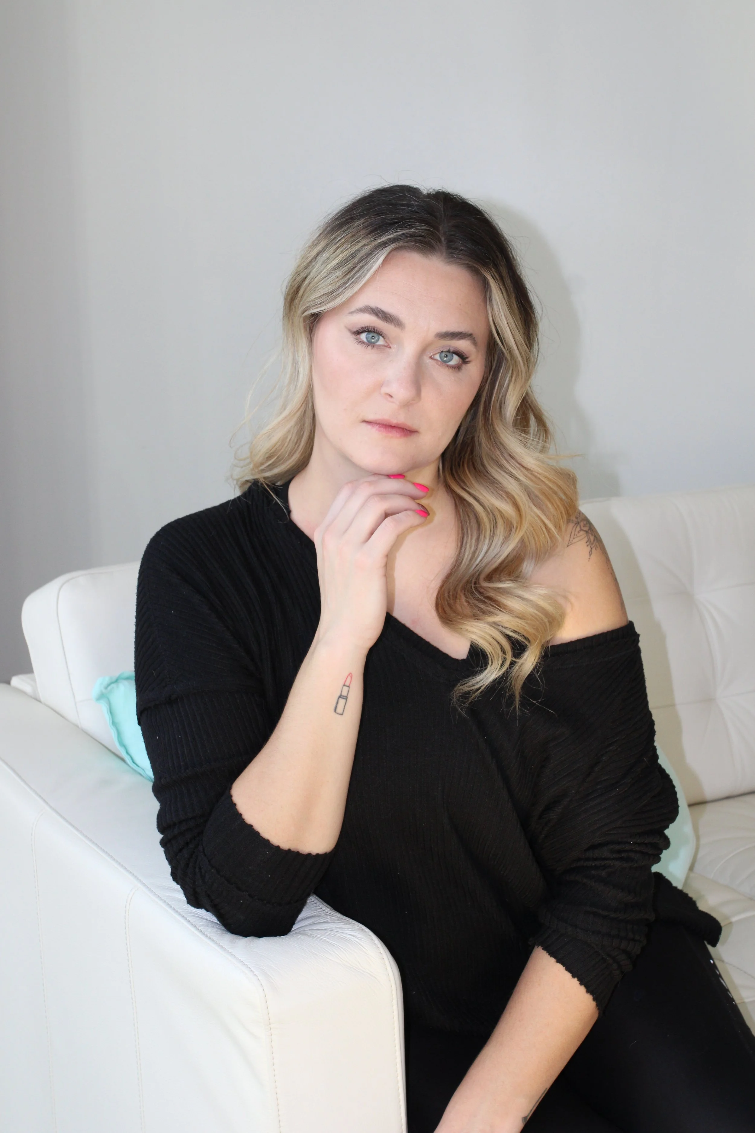 A woman with blonde hair and blue eyes sitting on a white couch, wearing a black top, with a hand near her chin and a small lipstick tattoo on her arm.