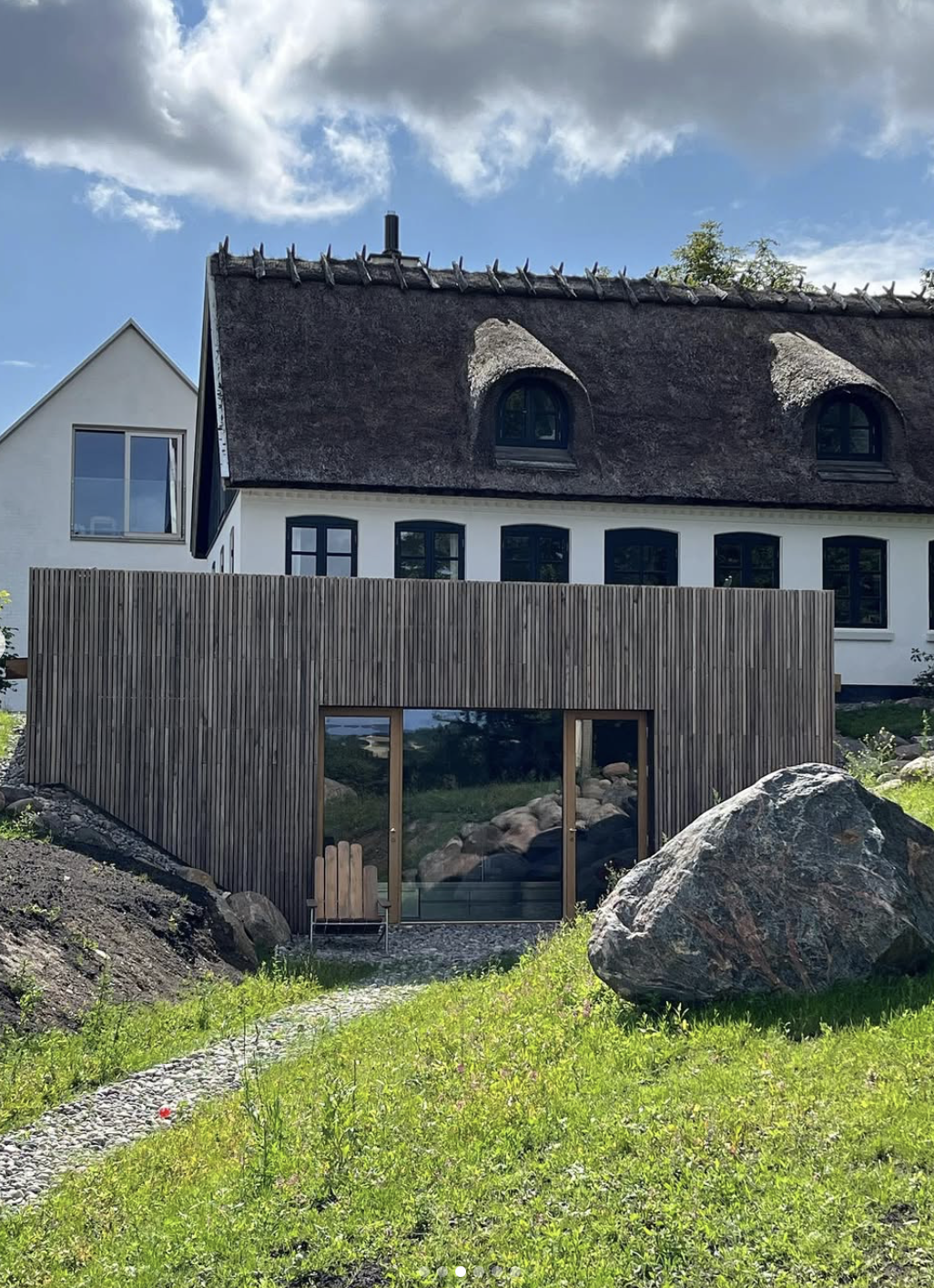 Et traditionelt stråtækt hus med moderne tilbygning i træ, omkranset af en frodig have med stor sten og græs.