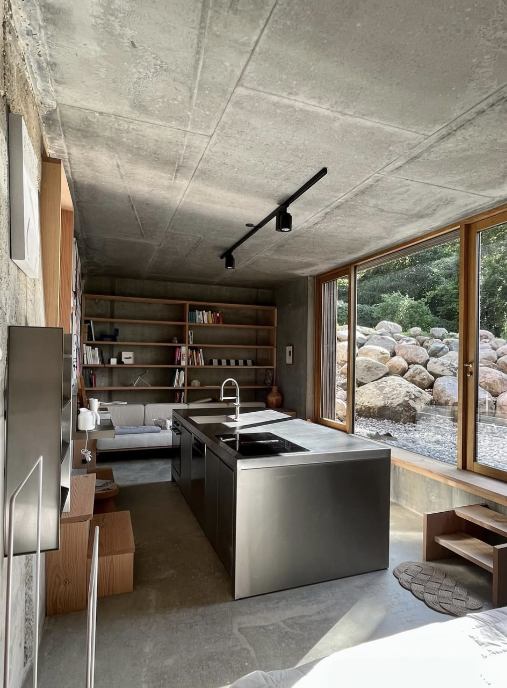 Moderne køkken med betonloft, stål køkkenø, store vinduer med udsigt til stenhave og indbyggede bogreoler.