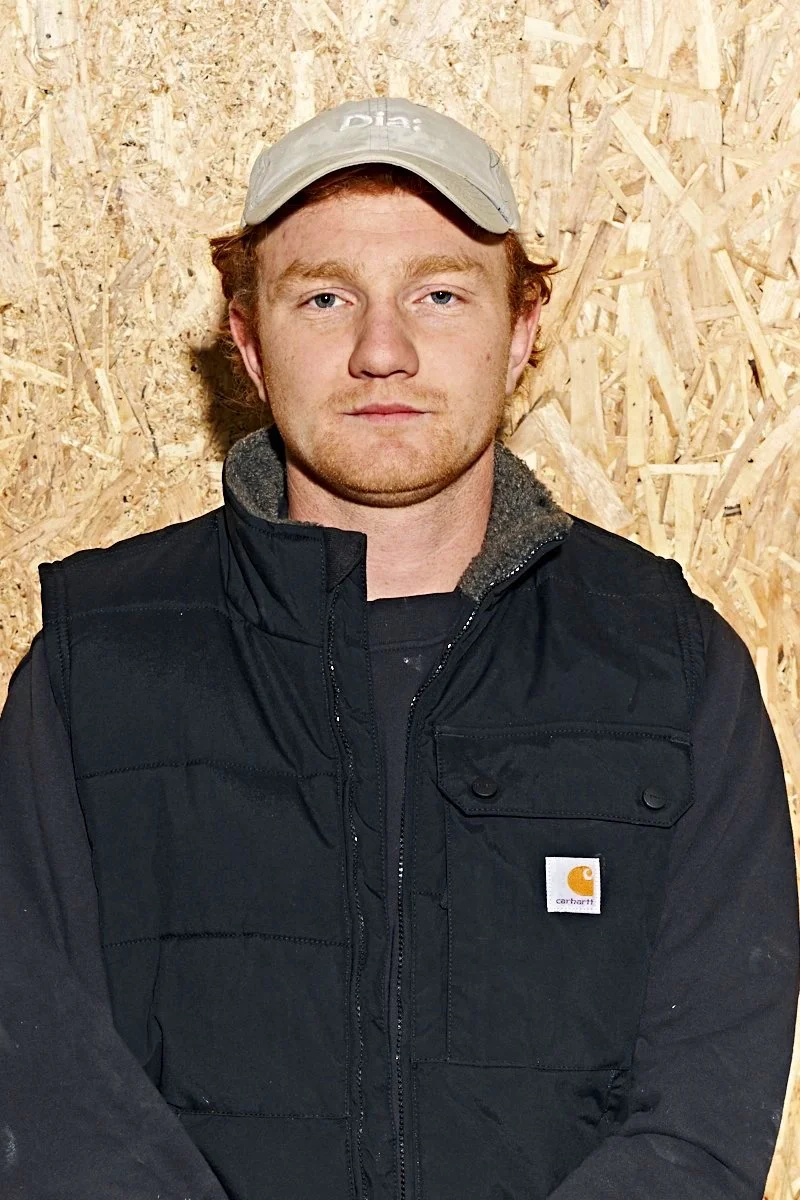 Mand med kasket og sort vest foran en trævæg.