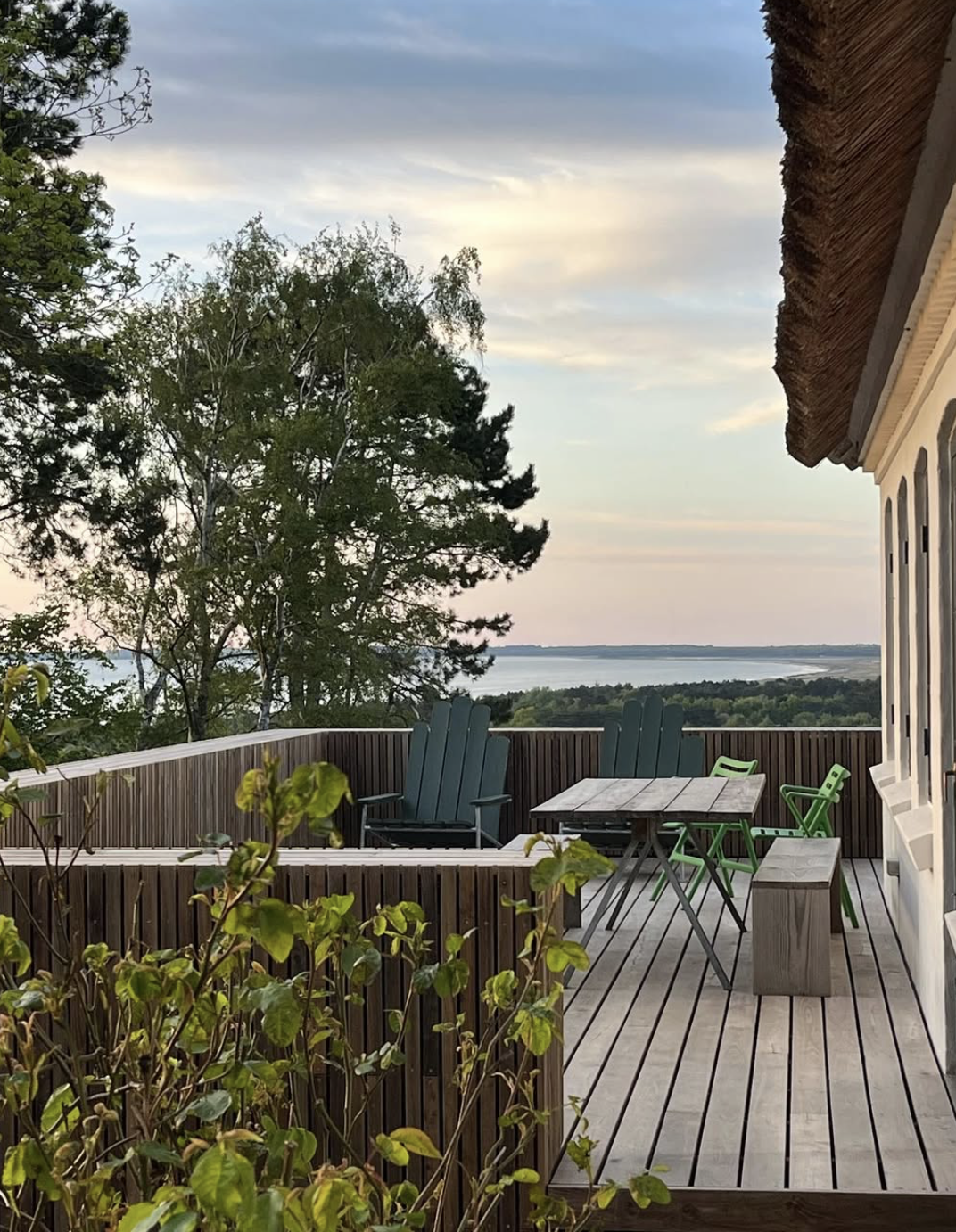 Træterrasse med bord og stole, omgivet af vegetation, udsigt til havet i baggrunden, solnedgangshimmel.