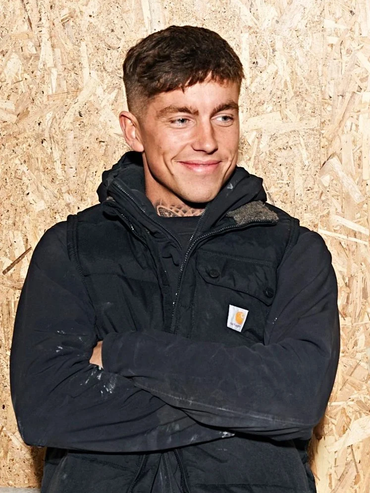 Mand i sort jakke står foran en krydsfinervæg og smiler.