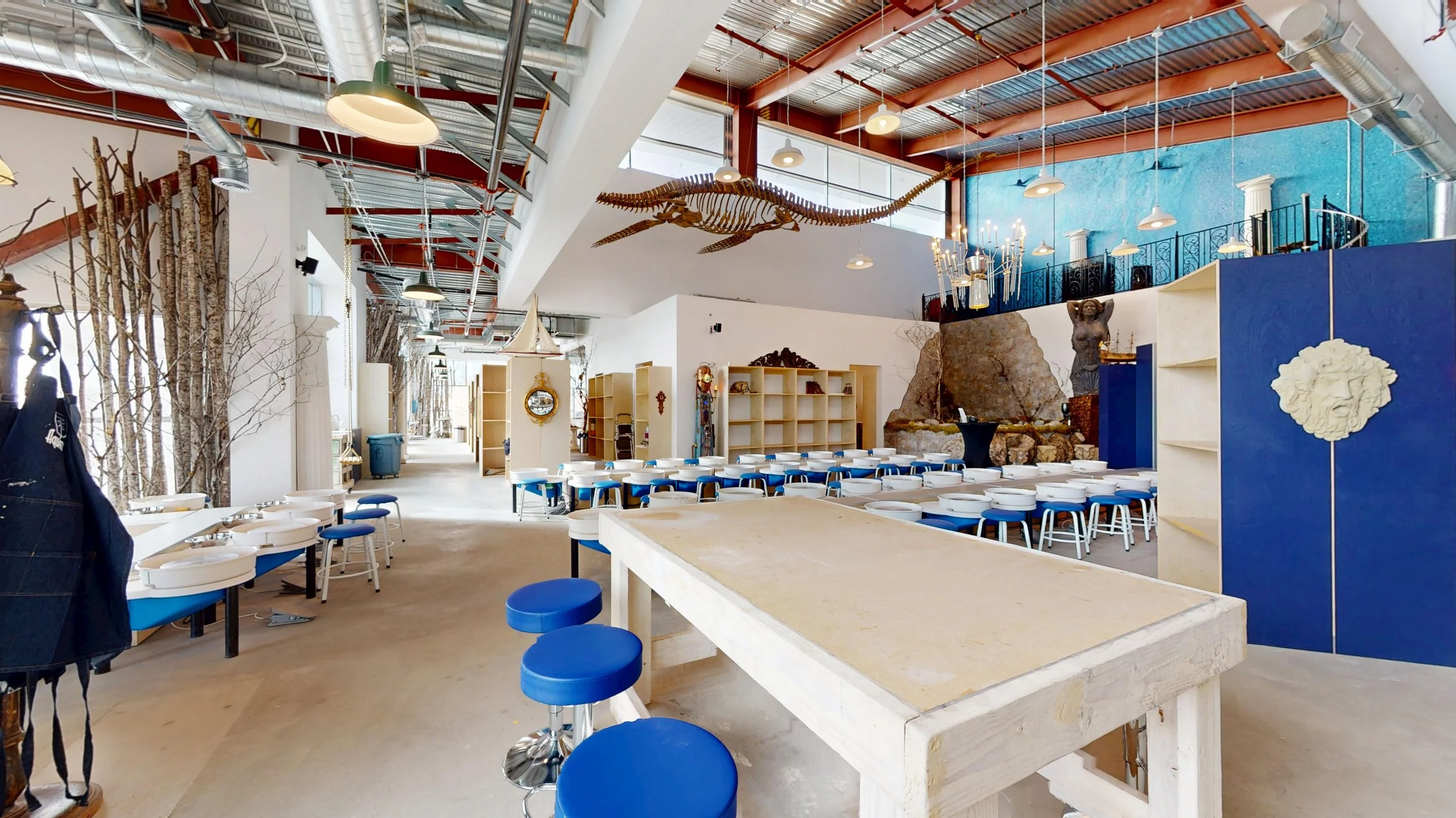 Interior view of a spacious restaurant with white and blue decor, wooden tables, and stools, featuring wall decorations including a large skeleton dinosaur, a sculpture of a woman, and various art pieces, with trees along the side walls.