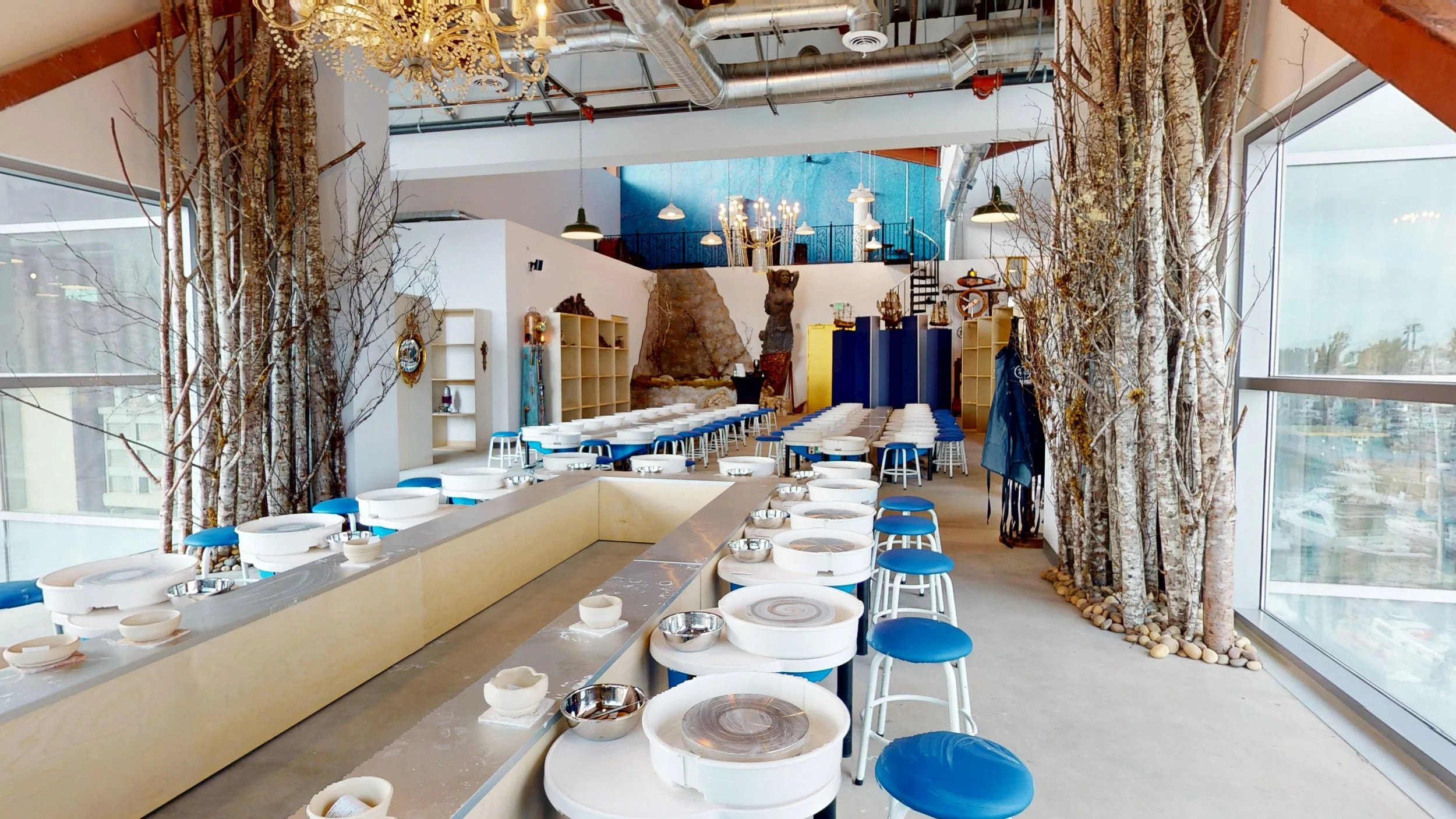 Interior of a pottery painting studio with pottery wheels, stools, and decorative trees. Large windows on the right let in natural light, and there is a chandelier hanging from the ceiling.
