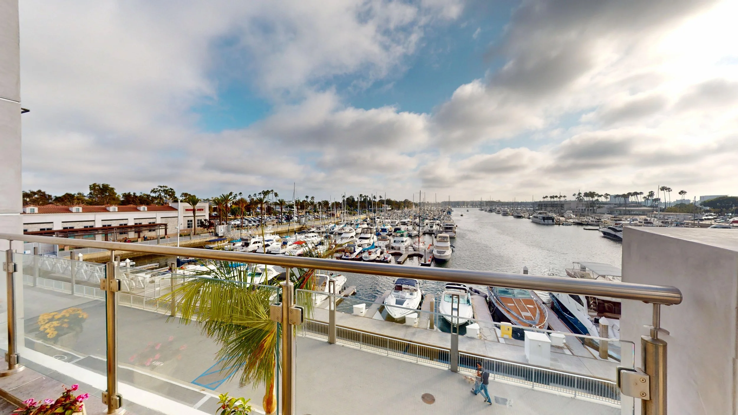View of a marina with numerous yachts and boats docked, seen from a balcony with glass railing. The sky is partly cloudy with blue patches, and there are palm trees in the background.