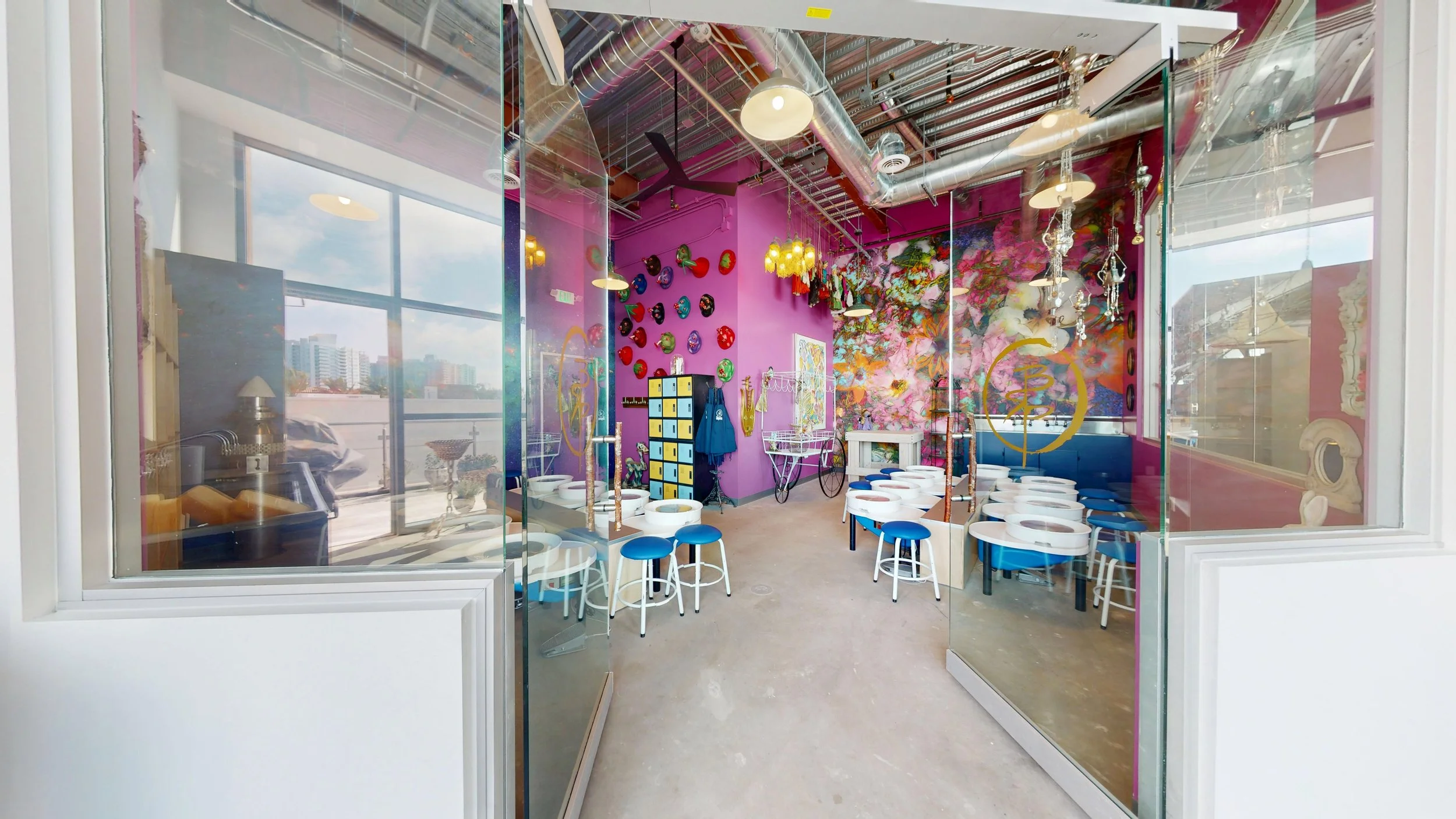 Colorful, modern cafe interior with vibrant pink walls, decorated with colorful masks, art, and hanging ornaments. White tables with blue and white chairs are arranged around the space, and large glass windows let in natural light. The ceiling featur