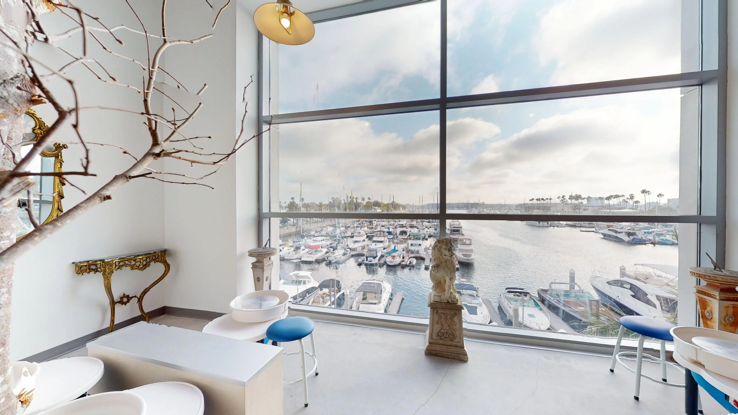 View of a marina through large floor-to-ceiling windows, showing boats docked in the water with palm trees in the distance, in a room with decorative furniture and sculptures.