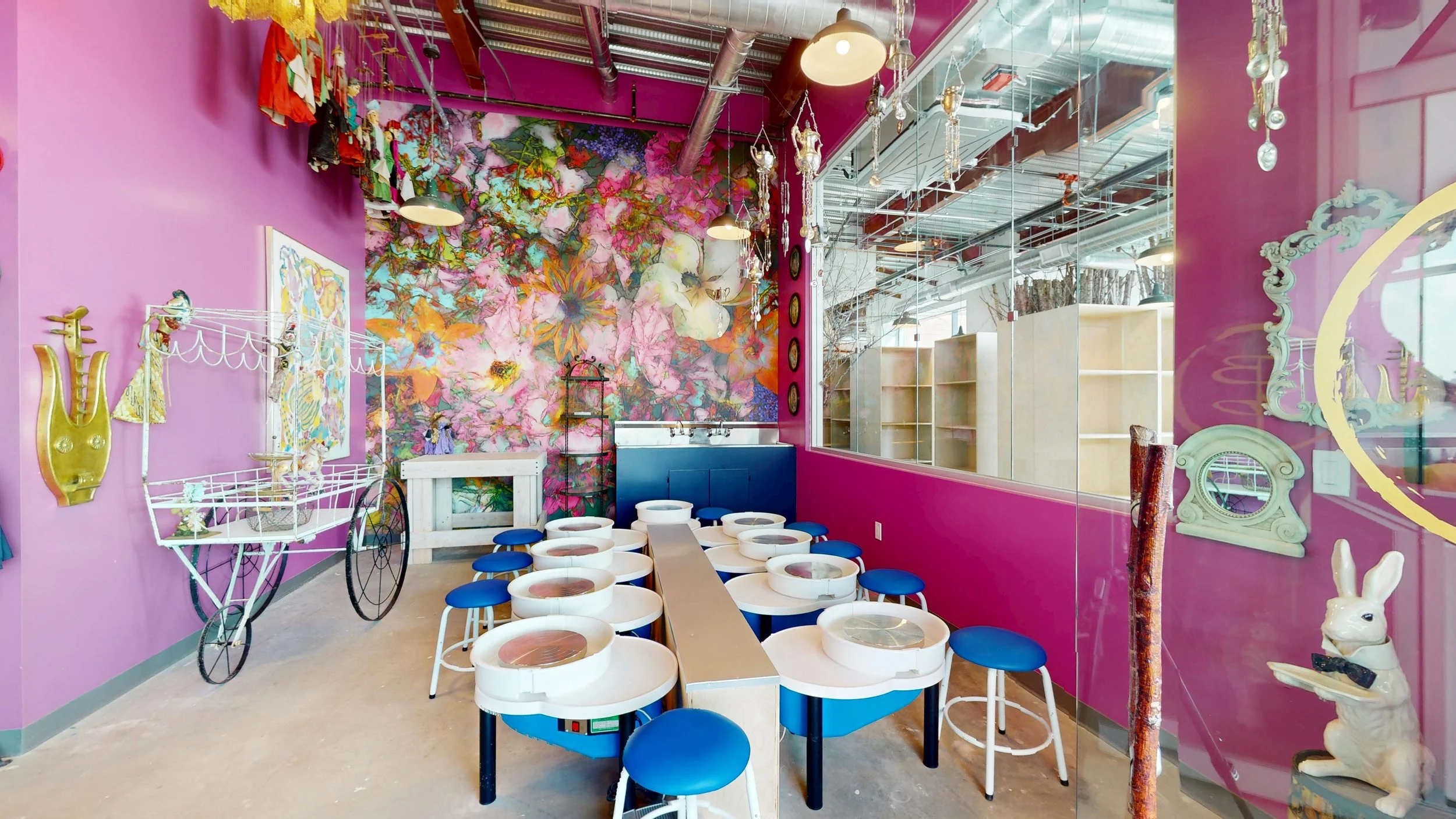 Colorful cafe interior with pink walls, floral mural, eclectic decorations including rabbit sculpture, vintage bicycle, ornate mirror, and modern seating with blue stools around pottery wheels.