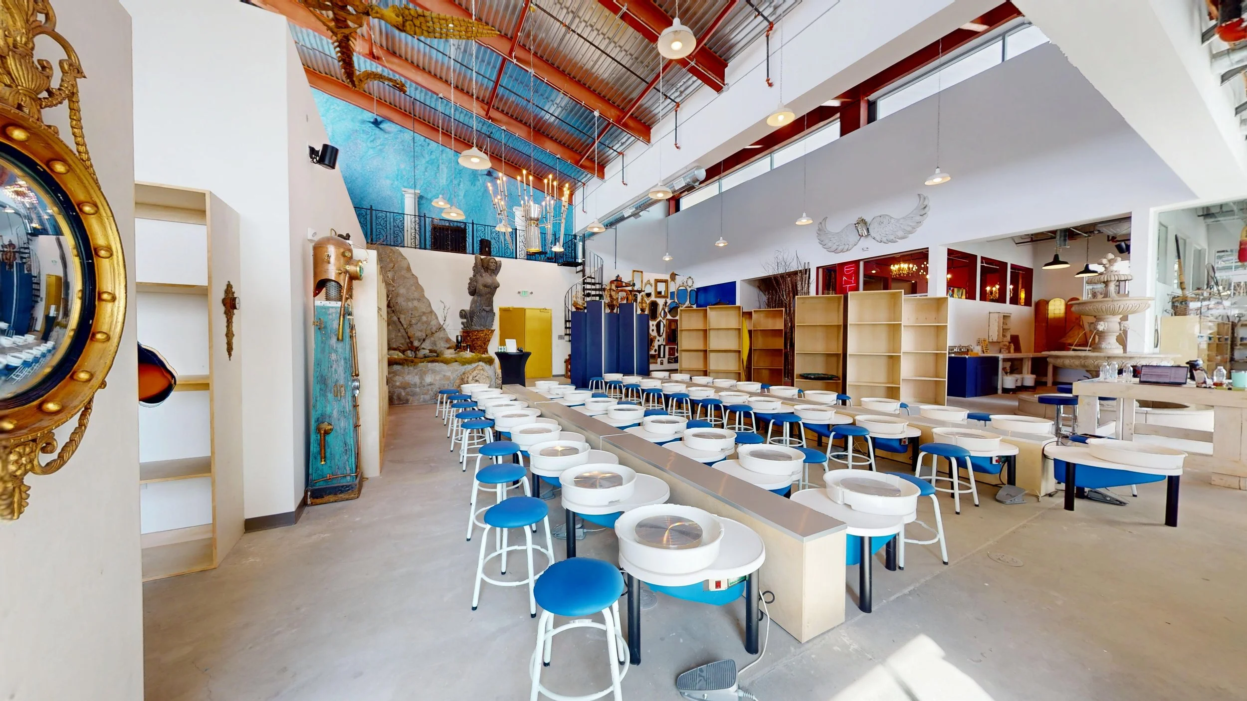Interior of an art gallery or store with empty display cases, blue stools, decorative sculptures, and wall art including a large blue mural and an ornate mirror.
