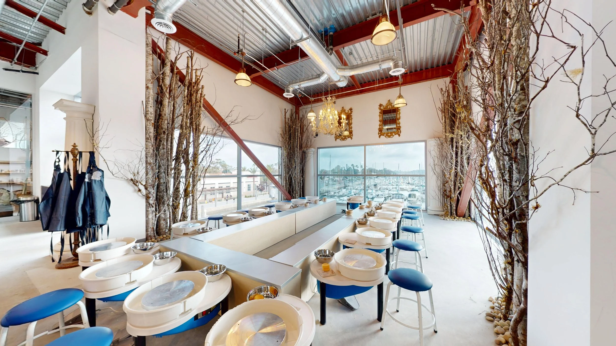 Interior view of a pottery painting studio with windows overlooking a marina, featuring white pottery wheels, blue stools, decorative tree branches, and mirrors on the wall.